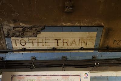 Hidden signs of Green Park station Photo of a cream tiled panel with blue border; TO THE TRAINS is painted on it. The tiles in the top left corner have broken away.