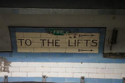 Hidden signs of Green Park station On a cream-and-blue tiled background, a cream tiled panel edged in blude says 'TO THE TRAINS' with arrows above and below the text. A tattered 'fire exit' sticker is on the top of the panel.