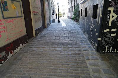 Photo of a small Parisian passage; in the foreground, the roadway is paved with stone around the edges and smaller, brown blocks in the middle. In the background is a charming pasage lined with buildings and with an old-fashioned lamp post.
