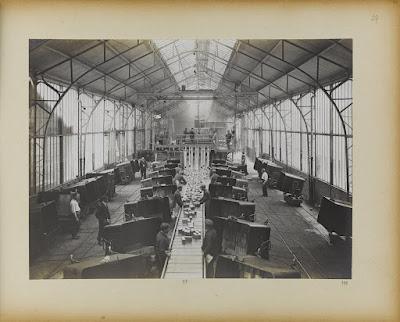 Vintage photo of a conveyor belt with wooden blocks on it. Either side, workers pack the blocks into wagonettes.