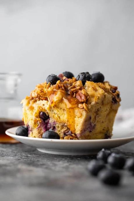 15+ Holiday Slow Cooker Recipes side view of a piece of french toast casserole on a white plate with blueberries on top and syrup behind it