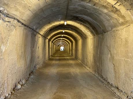 A long, dimly lit concrete tunnel inside Bunk’Art 1 in Tirana, Albania. The arched ceiling and rough walls lead toward a distant light at the end where two people are faintly visible.