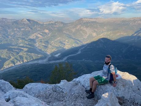 Alex Tiffany sitting on a white limestone rock near the summit of Mount Tujanit with vast mountain valleys stretching into the distance. He's enjoying the rewarding view after the steep climb above Tirana.