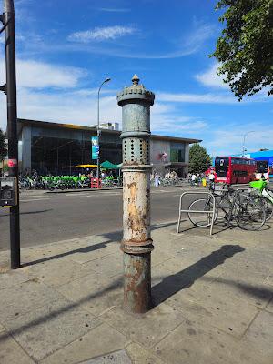 Photo of a metal pipe with decorative ridges, topped with a circle of crenellations and a spike. It has faded, rusting blue-and-white paint. About two-thirds of the way up, it is circled with holes in columns of six.