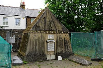 Photo showing a small building shaped like a rectangular tent with pitched roof, in - rather grimy - sandstone. To the sides are green netting erected by conservation workers, and in the background is a terrace of houses.