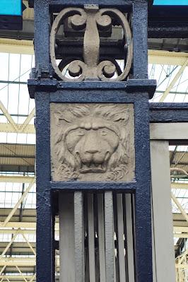 Photo of a blue metal square pillar, with beige metal details and a gold-brown inset relief of a lion's head.