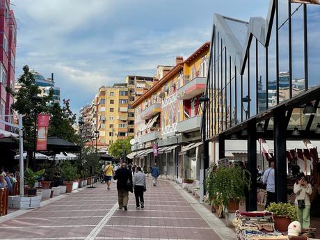 The lively Pazari i Ri (New Bazaar) market area in Tirana, Albania, with colorful buildings, glass-fronted market stalls, and locals enjoying the bustling atmosphere. People walk along the pedestrian street lined with plants, textiles, and cafes.