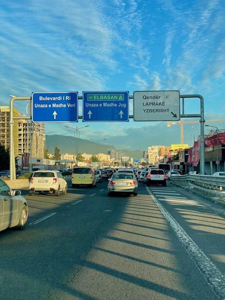 A busy highway in Tirana, Albania, during golden hour, showing traffic congestion and road signs directing to Bulevardi i Ri, Elbasan, and the city center. Construction cranes and buildings are visible in the background under a blue sky.