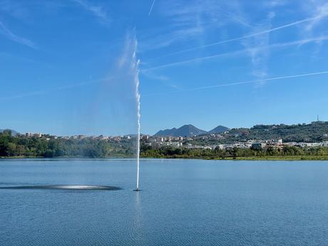 A fountain shoots a tall stream of water into the air at Tirana’s Artificial Lake, surrounded by greenery and distant buildings. The blue sky and scattered clouds add to the serene atmosphere.