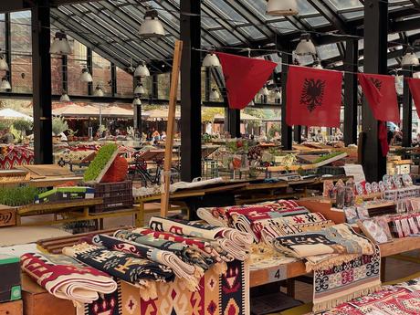 Inside Tirana’s Pazari i Ri (New Bazaar), a vibrant marketplace filled with traditional Albanian textiles, fresh produce, and artisanal goods. Red Albanian flags with the black double-headed eagle hang from the ceiling, adding a patriotic touch.