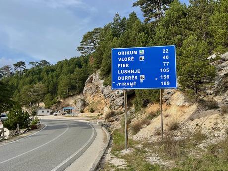 Road sign in southern Albania showing distances to Orikum, Vlorë, Fier, Lushnje, Durrës, and Tiranë, set against a backdrop of pine-covered hills. A clear view of Albania’s scenic mountain roads and well-marked driving routes.