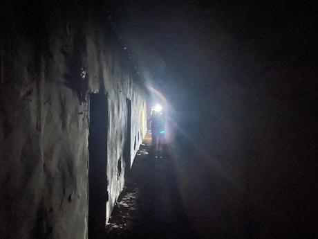 A dimly lit underground tunnel with rough concrete walls and narrow doorways, illuminated by a person’s flashlight at the far end. The light creates long shadows along the uneven floor and textured walls.