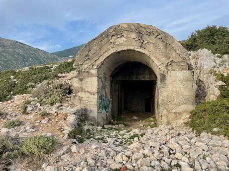The entrance of a large, weathered concrete bunker built into a rocky hillside along the Llogara Pass in Albania. Graffiti marks the archway, and rugged terrain with low shrubs surrounds the structure.