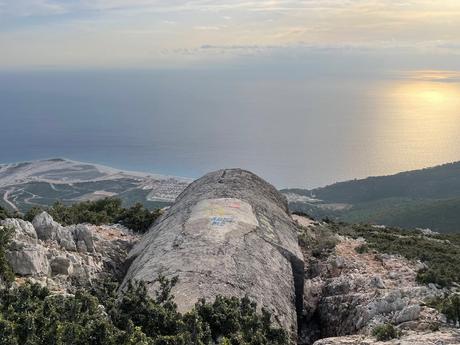A massive concrete bunker dome partly buried in rocky terrain overlooking the Albanian coastline. The sunlight reflects off the calm sea below, with the sky softly fading toward evening.