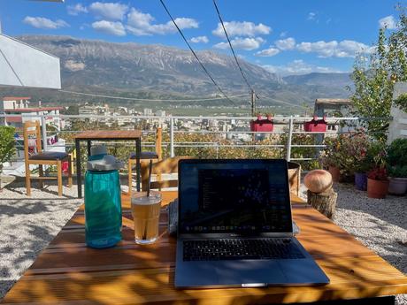 Outdoor workspace with a laptop, iced coffee, and water bottle on a wooden table overlooking Gjirokaster’s valley and mountain views. A relaxing travel moment that showcases why working or relaxing with a view is among the best things to do in Gjirokaster.