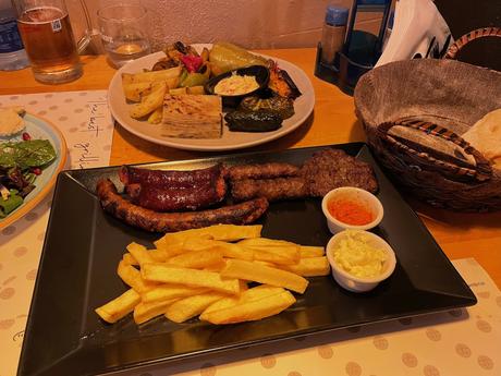 Traditional Albanian mixed grill served with chips, red pepper sauce, and garlic dip, alongside roasted vegetables and bread. A hearty Balkan-style meal showcasing Albania’s love of grilled meats and homely flavours.