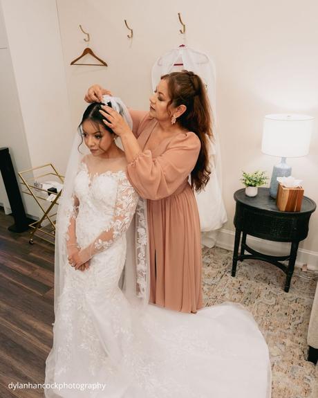 An Emerald & Sage, Zero-Alcohol Real Wedding a bride in a white lace gown sits on a plush rug as her mother gently adjusts her veil dylanhancockphotography