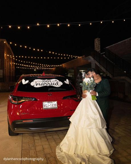 An Emerald & Sage, Zero-Alcohol Real Wedding a couple shares an intimate kiss in front of their Just Married decorated car under the warm glow of string lights at night dylanhancockphotography