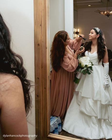 An Emerald & Sage, Zero-Alcohol Real Wedding a bride in a white gown and gloves stands before a large wooden framed mirror as a mom beside her applies makeup dylanhancockphotography