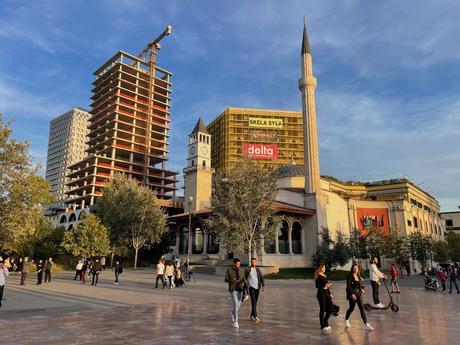 Is Albania Safe To Visit? Skanderbeg Square in Tirana, Albania, during sunset, featuring the Et'hem Bey Mosque, the Clock Tower, and a mix of modern and historical architecture. People are strolling across the square, and a construction crane looms in the background.