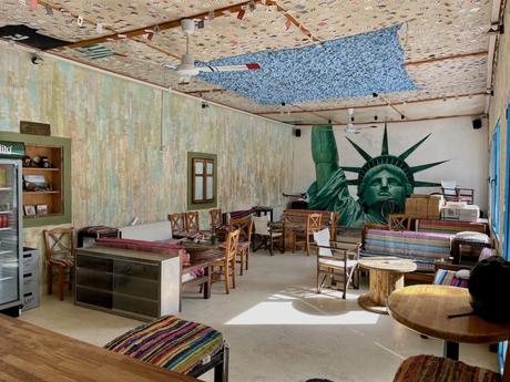 Inside a colourful cafe with cozy furniture, a prominent mural of the Statue of Liberty adds a striking visual to the room. The ceiling is decorated with various patterns and papers.