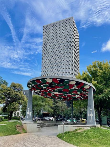 A modern high-rise building in Tirana, Albania, with a unique geometric facade, next to a colorful urban canopy made of red, green, and black circular panels. The vibrant sky and lush greenery frame the cityscape.