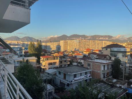 A scenic urban view from a balcony in Komuna e Parisit, Tirana, Albania, during golden hour. The foreground features a mix of residential buildings with flat and sloped roofs, some with laundry hanging outside. In the background, a large modern apartment complex and distant mountains bathed in warm sunlight contrast against the clear blue sky with a few wispy clouds.