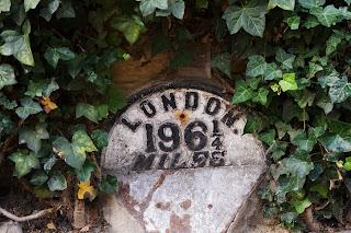 Small milestone with a curved top, painted white with black lettering and surrounded by ivy. It says that London is 196 and a quarter miles.