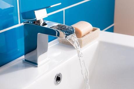 Close-up image of a modern chrome mixer tap with water flowing into the sink nearby soap in front of blue tiled wall