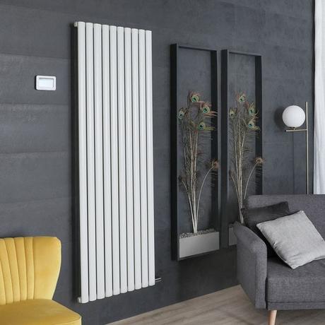 8 radiator ideas for period properties ECOSO Aruba Ardus white dry heat vertical electric designer radiator on dark wall nearby yellow chair and gray chair and two vertical black frames featuring peacock feathers