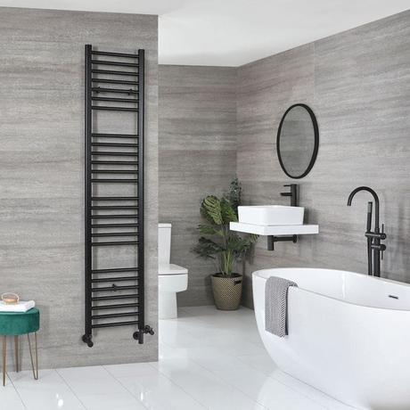 Milano Nero matt black dual fuel straight heated towel rail on a gray wall in a modern bathroom nearby white toilet, sink beneath a round black mirror and white freestanding bathtub