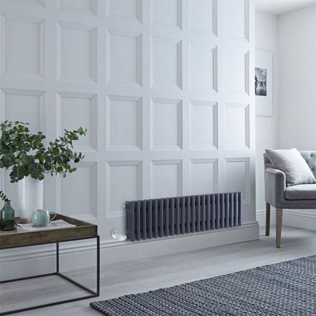 Milano Windsor anthracite traditional horizontal electric triple column radiator on white wall nearby gray chair and small brown table with white vase