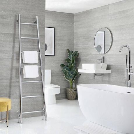 Milano Indus chrome floor-standing ladder heated towel rail leaning on gray wall nearby small yellow stool and large white freestanding bath in a bathroom also featuring a large houseplant and large circular mirror above a white sink on floating white shelf