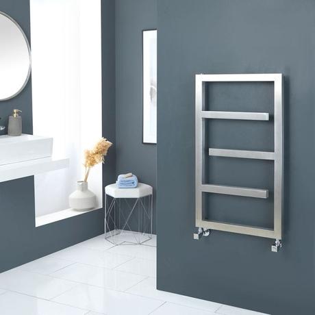 Milano Eclipse designer satin stainless steel heated towel rail on dark wall around the corner from small stool, vase and white sink on white shelf beneath a large circular mirror