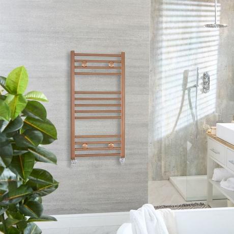 Milano Ive metallic copper straight heated towel rail on light gray wall nearby shower enclosure and floorstanding white vanity unit with white sink