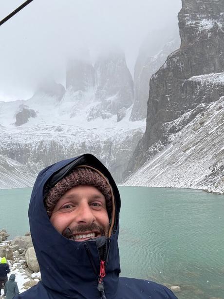 Alex Tiffany taking a selfie beside the turquoise lagoon and snowy cloud-covered towers at Mirador Base Torres
