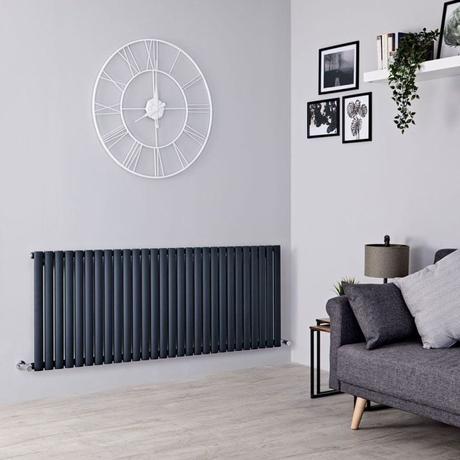 Milano Aruba black horizontal designer radiator on white wall beneath large clock nearby multiple framed pictures and shelving above gray settee