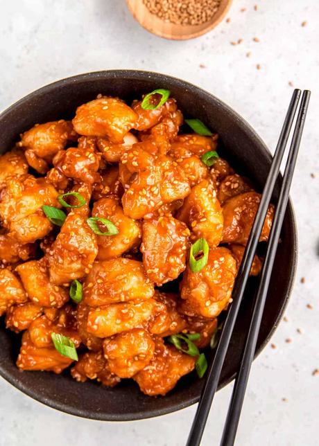 black bowl filled with sticky sesame chicken garnished with sesame seeds and green onions