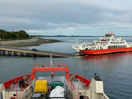 View from a vehicle ferry approaching a landing in southern Chile, with another red-and-white ferry docked beside the shore.
