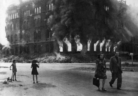 A German couple strolls past a burning building. Bizarre scenes like this were common at this time and place.